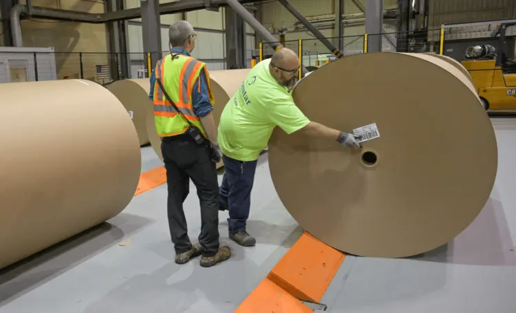 Two men in safety vests are handling large rolls of paper in a work environment.