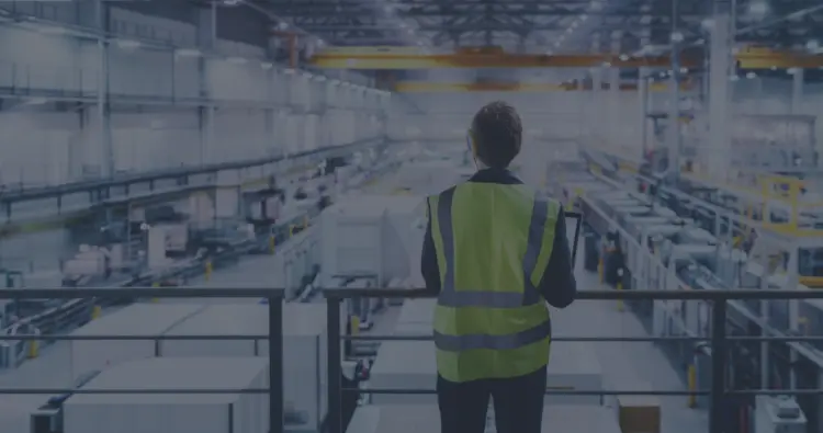 A man wearing a safety vest stands in front of a factory, evaluating its structure and environment.