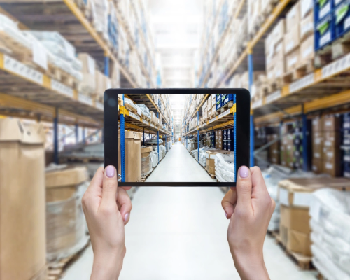 A person in a warehouse holds up a tablet, showcasing information or data to an unseen audience.